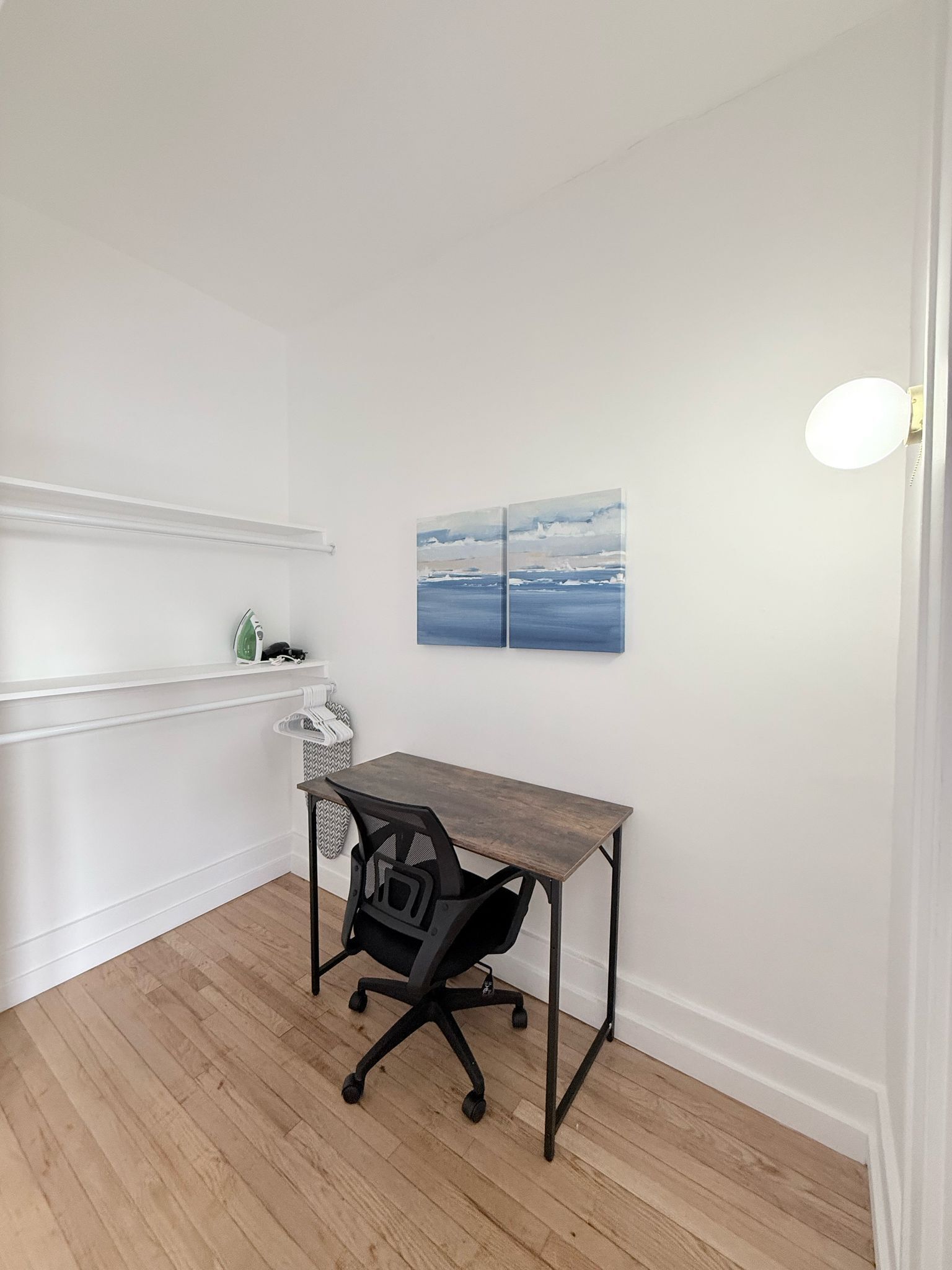 A small, neatly organized room with a wooden desk, a black office chair, and a wall-mounted shelf. There is a painting of a seascape above the desk.