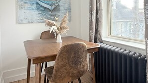 A dining area with a wooden table, a chair, a painting of a boat, a vase with dried plants, and a window with curtains. - Modern studio in Downtown Rochester with a full kitchen and Smart TV, steps from High Falls. (Rochester)