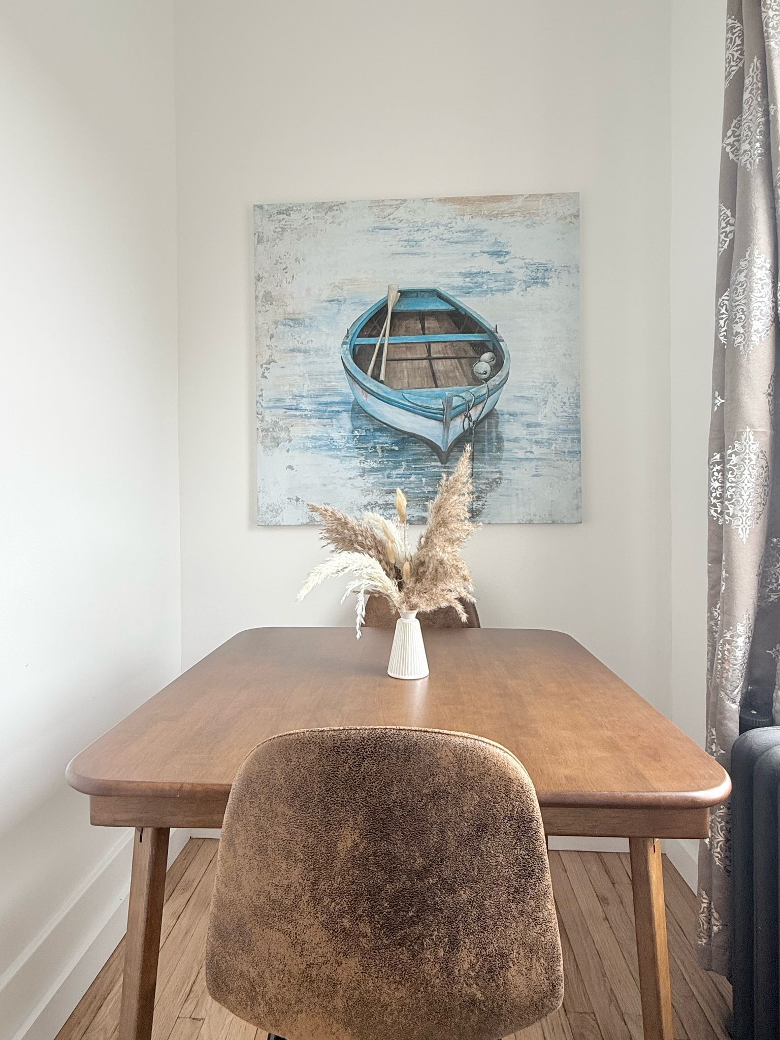 Een eethoek met een houten tafel, een stoel, een schilderij van een boot en pampa's in een vaas.