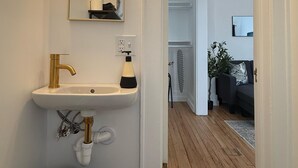 Ett badrum med ett vitt handfat, en guldgul kran och en spegel ovanför. På golvet ligger en svart hatt och det står en vit papperskorg där.
