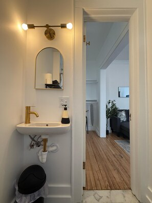 A bathroom with a white sink, gold faucet, and a mirror above it. There is a black hat on the floor and a white trash bin. - Stylish High Falls studio with a kitchen, Smart TV, and prime downtown location. (Rochester)