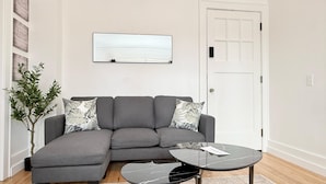 Ett modernt vardagsrum med en grå hörnsoffa, ett soffbord med glasskiva, en krukväxt och en platt-TV som är monterad på väggen.