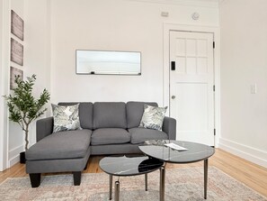 A modern living room with a grey sectional sofa, a glass-top coffee table, a potted plant, and a flat-screen TV mounted on the wall. - Stylish High Falls studio with a kitchen, Smart TV, and prime downtown location. (Rochester)