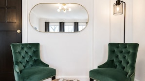 A room with two green upholstered chairs, a round wooden table with a book, a large mirror, and a floor lamp. - Studio Near Downtown+Wi-Fi+Queenbd_HighFall. (Rochester)
