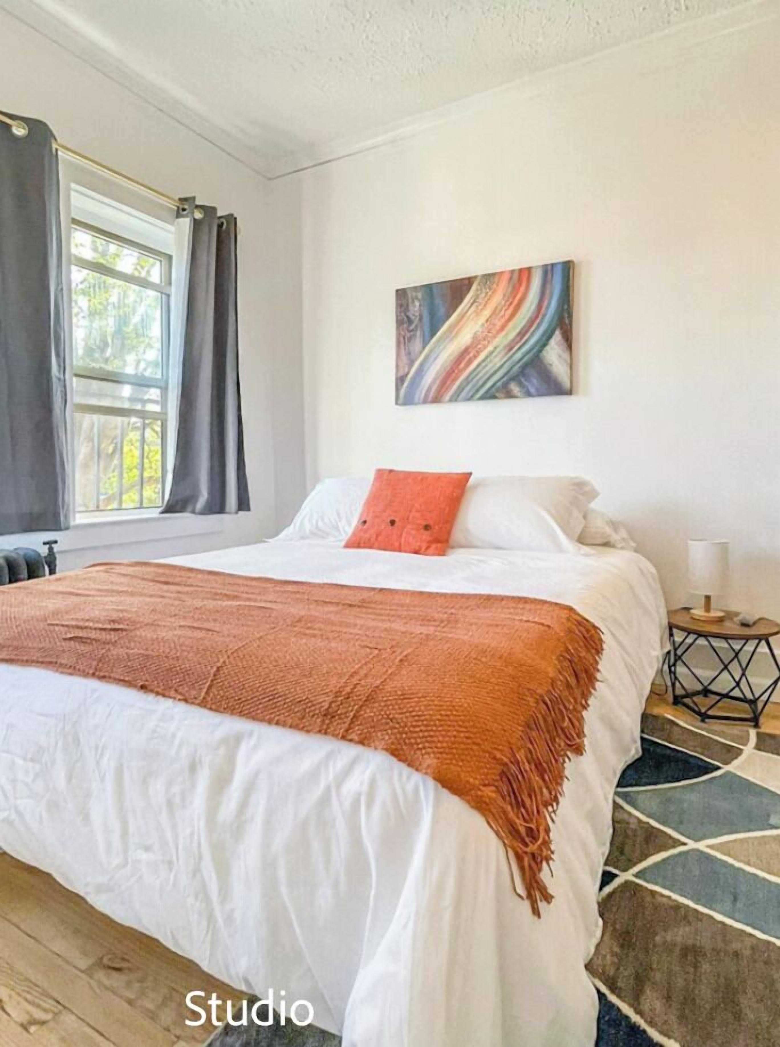 A bedroom with a large bed, a window with curtains, a painting on the wall, and a small bedside table.