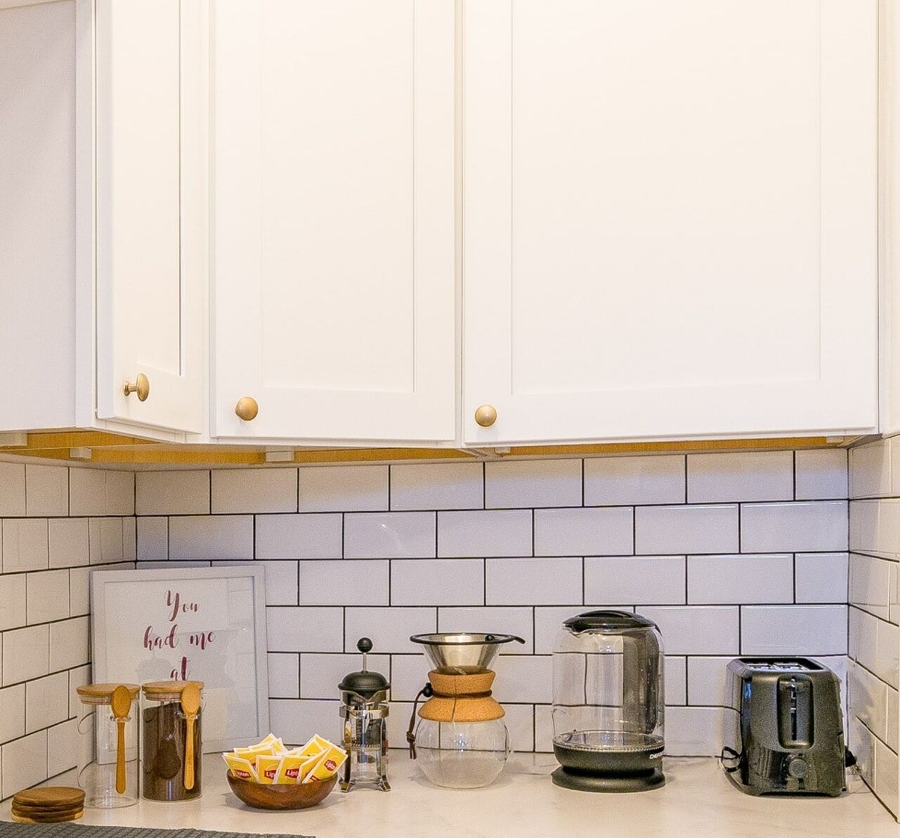 A kitchen with white cabinets, a tiled backsplash, a coffee maker, a glass carafe, a toaster, and a sign that reads "You had me at."