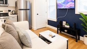 Ett modernt vardagsrum med en vit soffa, ett soffbord, en platt-TV som är monterad på en mörk vägg, ett köksområde med ett kylskåp i rostfritt stål och en mikrovågsugn.