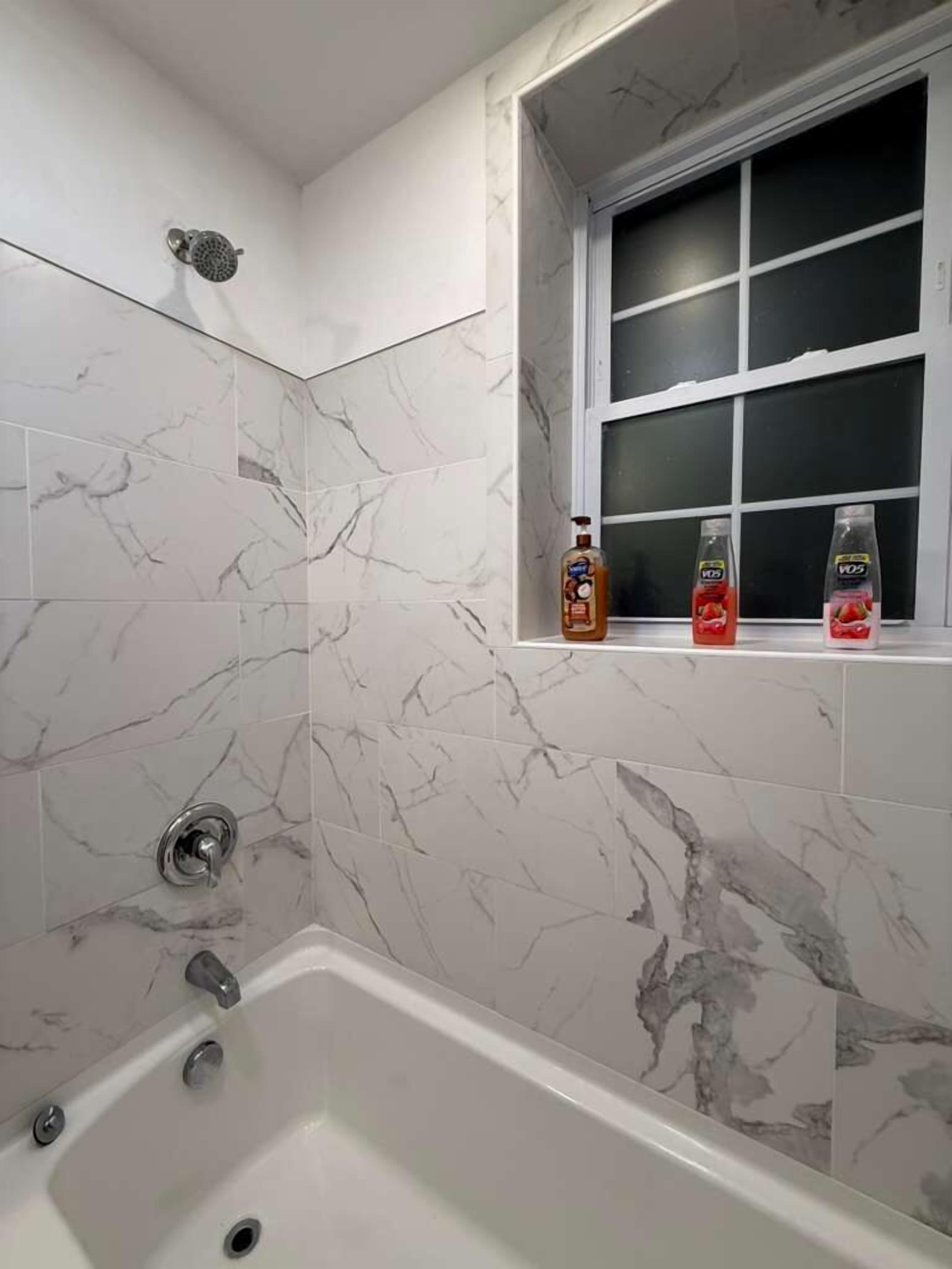 Un baño con zona de ducha de azulejos de mármol, una bañera y un ventanal con repisa que sostiene dos botellas.