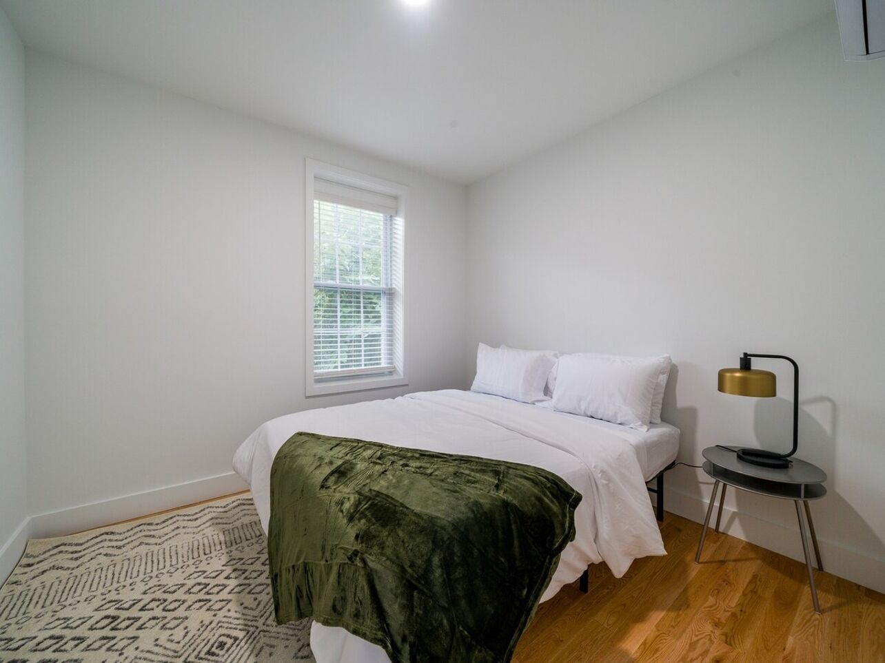 Un dormitorio con cama, lámpara de noche, ventana y alfombra estampada.