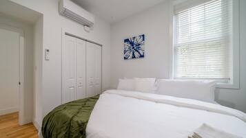 A bedroom with a large bed, white walls, a window with blinds, a door, and a wall-mounted air conditioner.