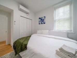 A bedroom with a large bed, white walls, a window with blinds, a door, and a wall-mounted air conditioner. - Cornhill 2BDRM-HighFall+EquippedKitchen+Wi-Fi (Rochester)
