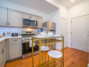 A compact kitchen with white cabinets, a stainless steel microwave, oven, and refrigerator. There is a small table with stools and a wine glass on the counter. - Cornhil 1Bed-EquippedKitchen,HighFalls,DT,TV_WiFi (Rochester)