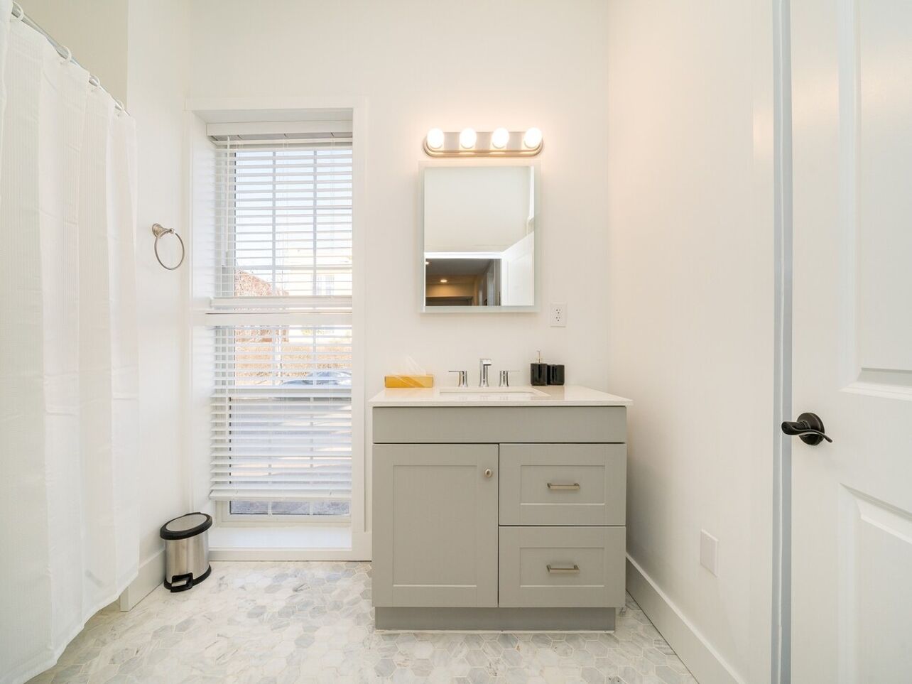 A bathroom with a vanity, a mirror with lights, a window with blinds, and a door.