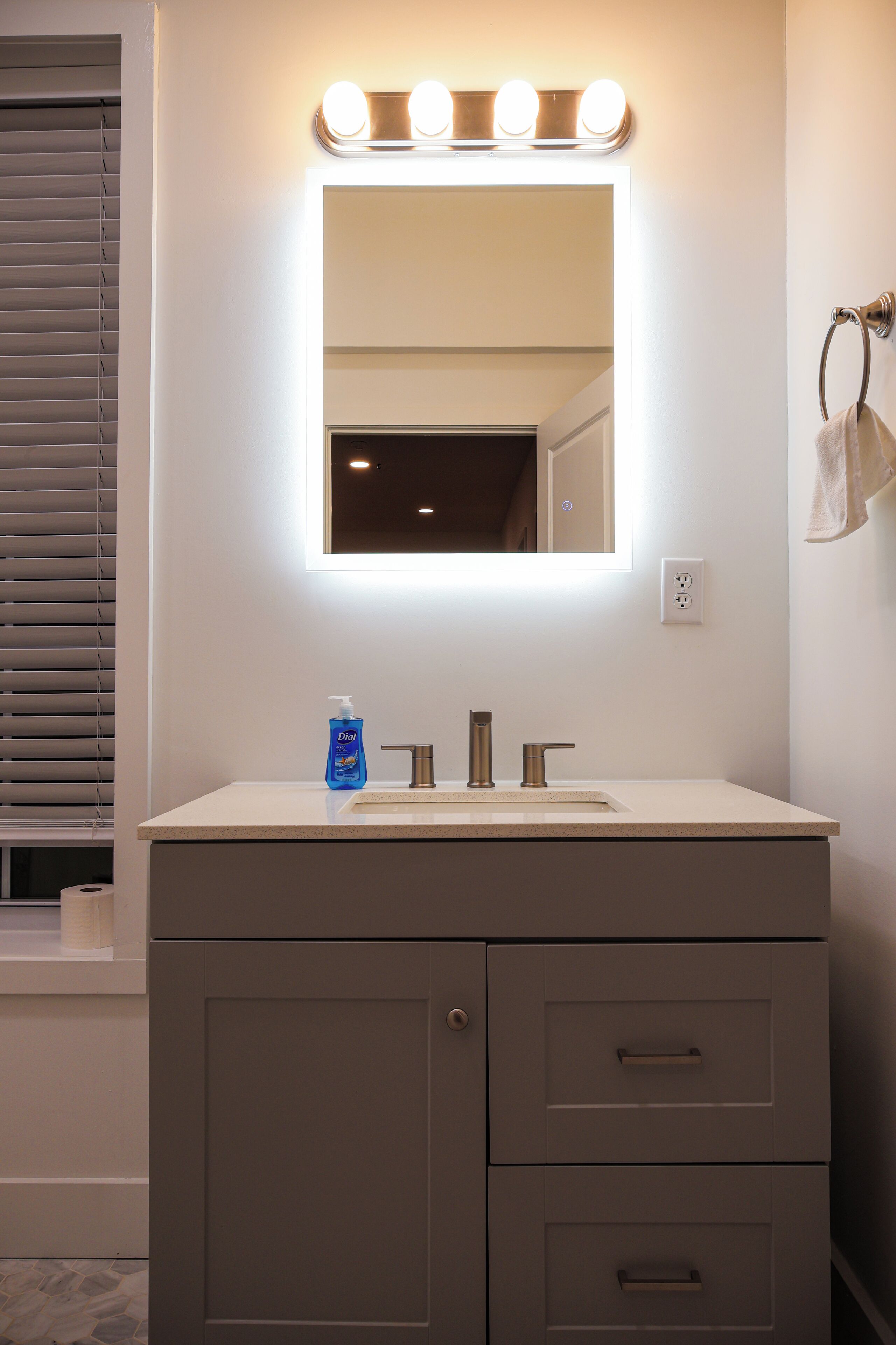 Un baño con tocador, espejo con iluminación, jabón de manos y toallero.