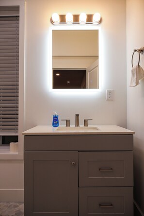 A bathroom with a vanity, a mirror with lights, a bottle of hand soap, and a towel rack. - 1BED+EquippedKitchen+HighFalls+TV+DT_Wi-fi (Rochester)