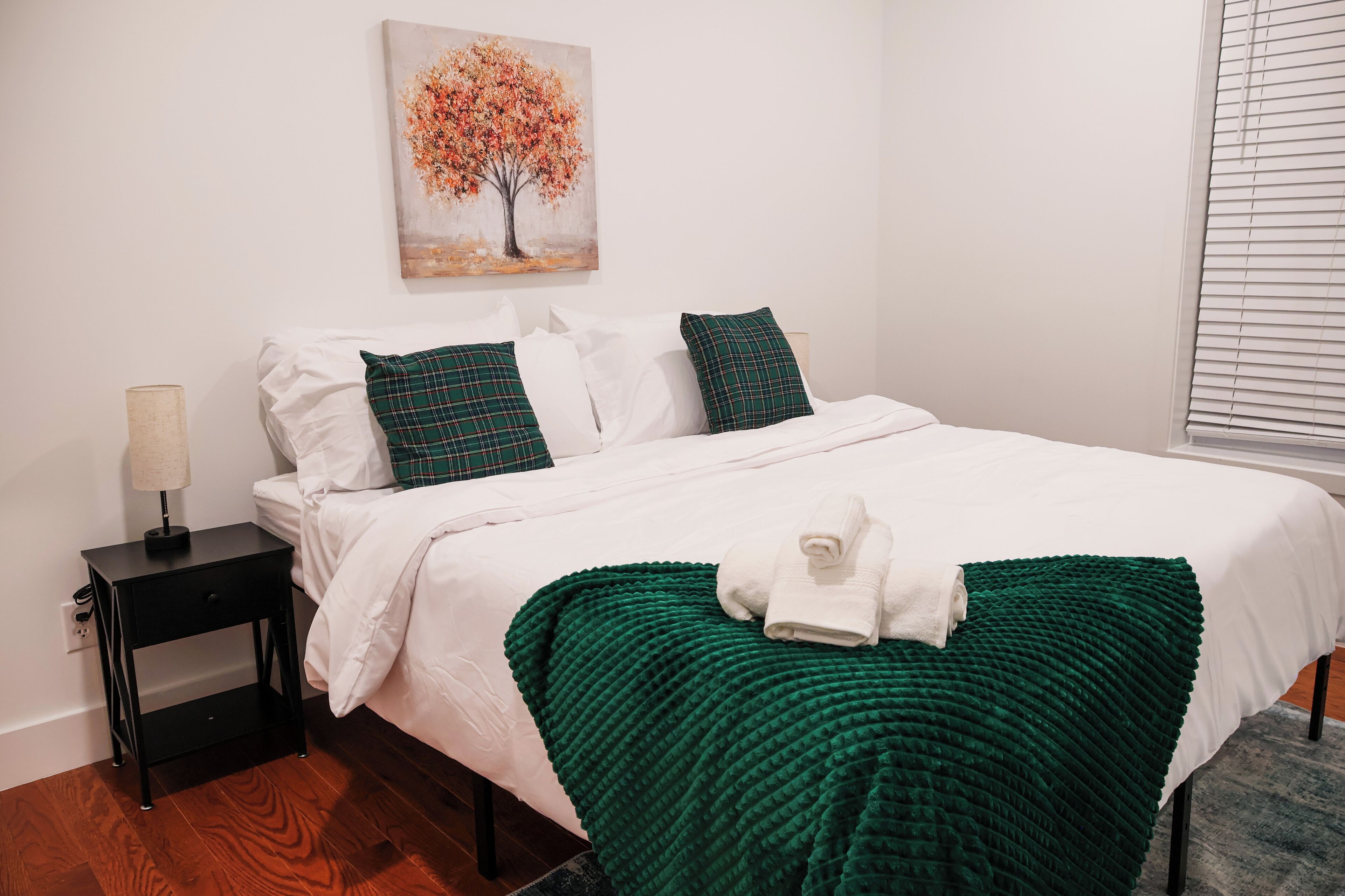 Un lit bien fait, avec du linge de lit blanc et des touches de vert, une table de chevet avec une lampe et un tableau représentant un arbre accroché au mur.