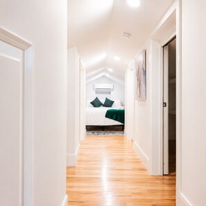 A narrow hallway with wooden flooring leading to a bedroom with a bed, pillows, and a green blanket.