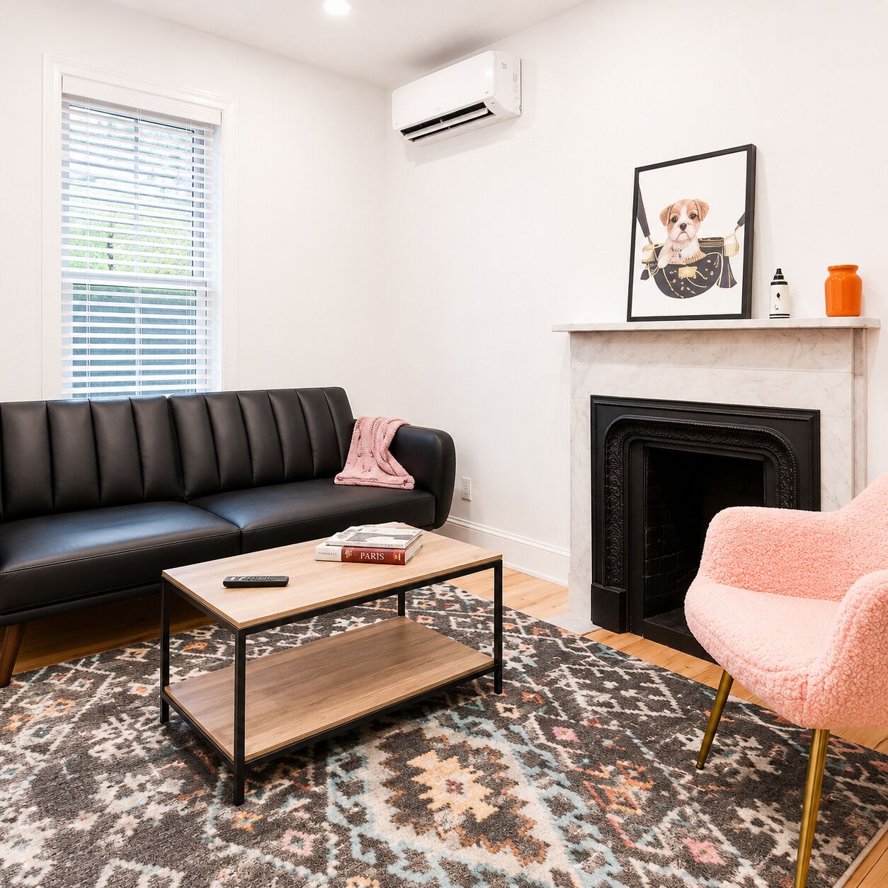 A living room with a black leather sofa, a wooden coffee table, a pink chair, a patterned rug, a fireplace, and an air conditioning unit.