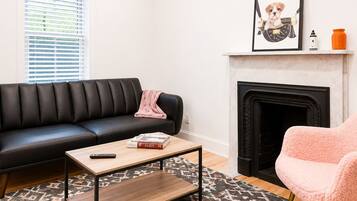 A living room with a black leather sofa, a wooden coffee table, a pink chair, a patterned rug, a fireplace, and an air conditioning unit.