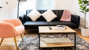 A living room with a black sofa, two white pillows, a pink chair, a wooden coffee table, a patterned rug, a potted plant, and a wall-mounted air conditioner. - Cozy 1BR with equipped kitchen, dining table, TV, Wi-Fi, near High Falls (Rochester)
