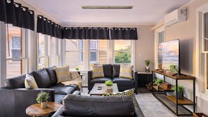 Ein modernes Wohnzimmer mit schwarzen Ledersofas, einem Couchtisch und einem Fernseher auf einem Holzständer. Große Fenster mit schwarzen Vorhängen lassen viel Tageslicht herein.