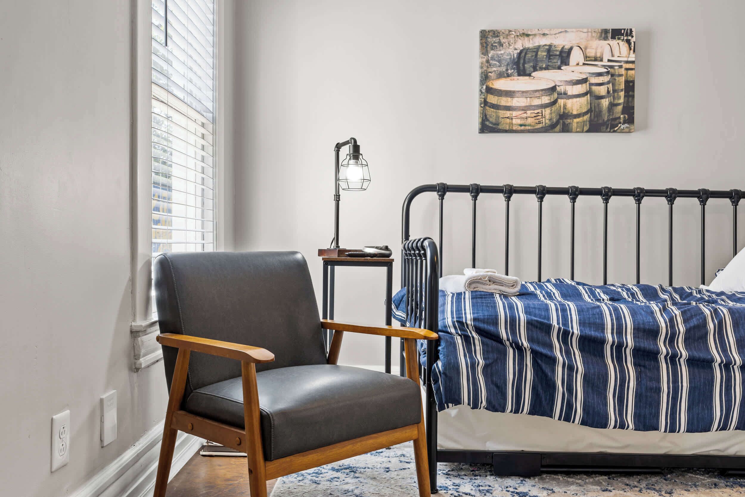 Quarto com cama, cadeira, mesa de cabeceira, abajur e janela com persianas.