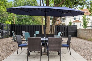 Eine Terrasse mit Tisch und Stühlen unter einem großen Sonnenschirm, umgeben von einem Holzzaun und Bäumen.