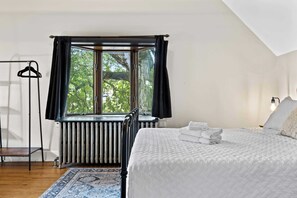 Una camera da letto con un letto, un termosifone, una finestra con tende e un appendiabiti, con vista su alberi.
