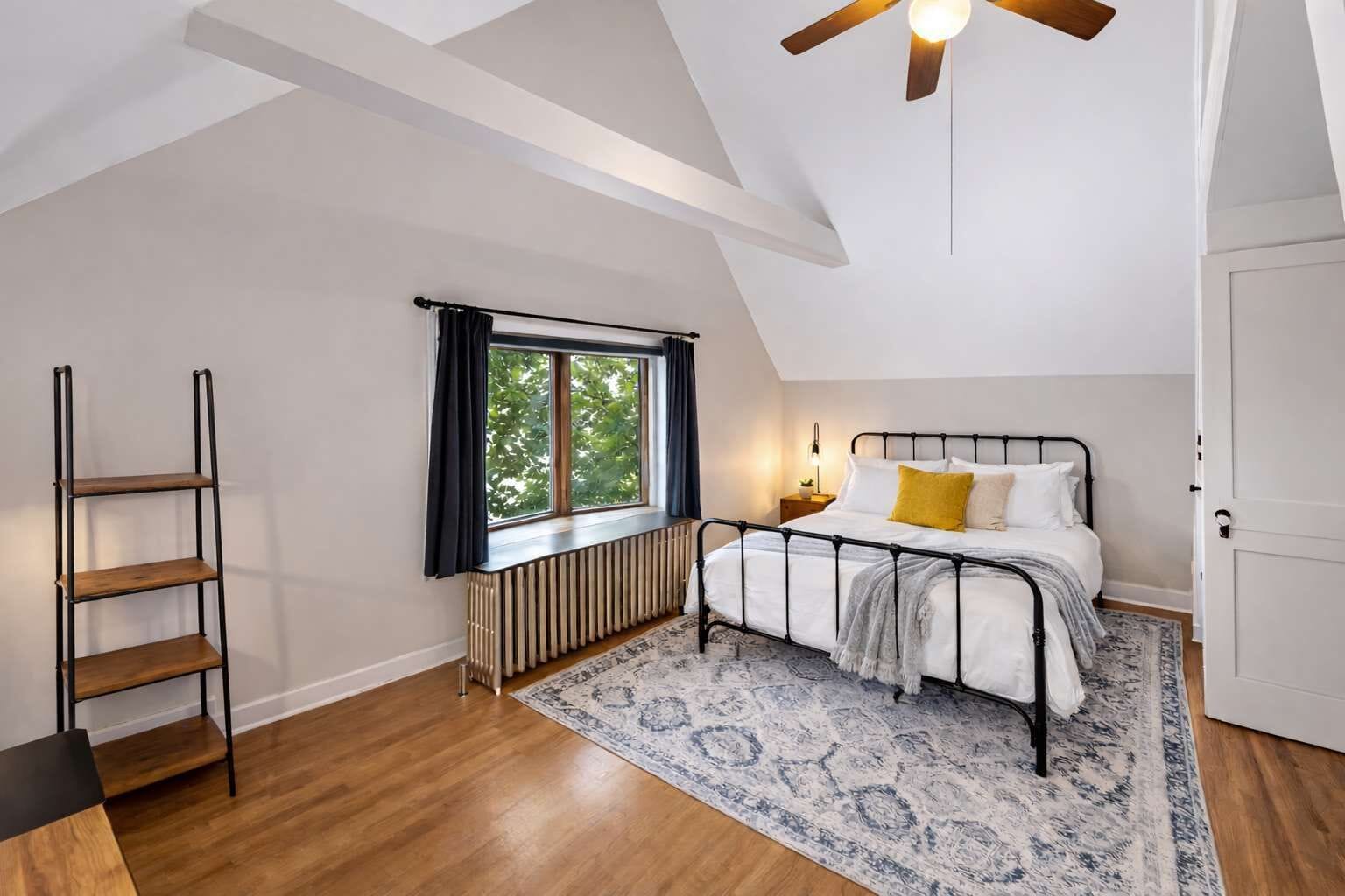 A bedroom with a loft-style ceiling, a bed with a black metal frame, a radiator, a wooden ladder, and a large window with curtains.