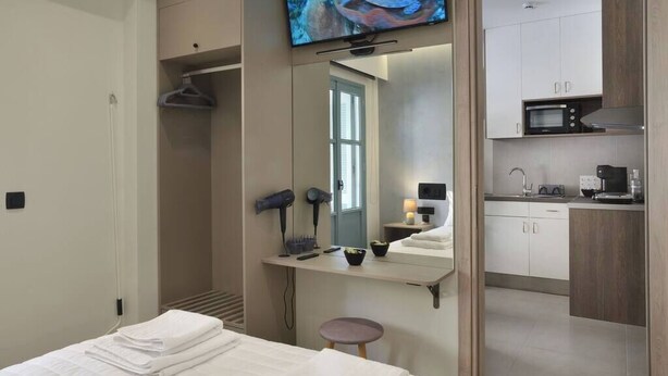 A hotel room with a bed, a TV, a mirror, a small table with a hairdryer, and a kitchenette. - Amara Porto Living | Apartment 1 (Chania)