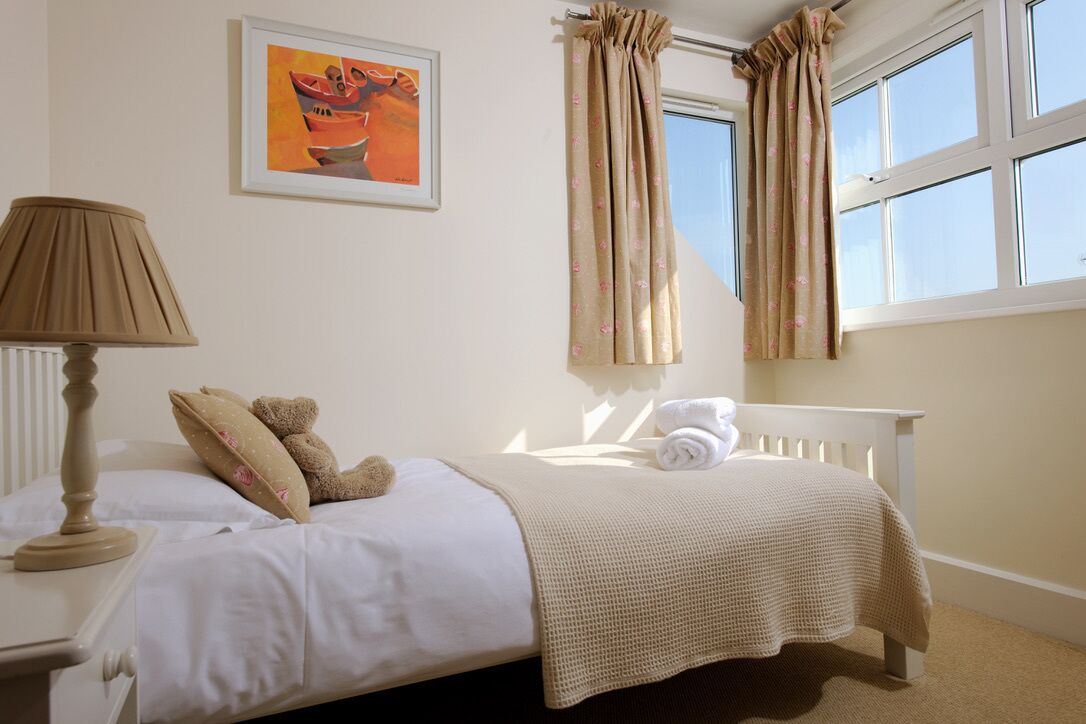 A neatly made bed with a white comforter and a beige knitted blanket, a bedside lamp, a teddy bear, and a framed artwork on the wall.