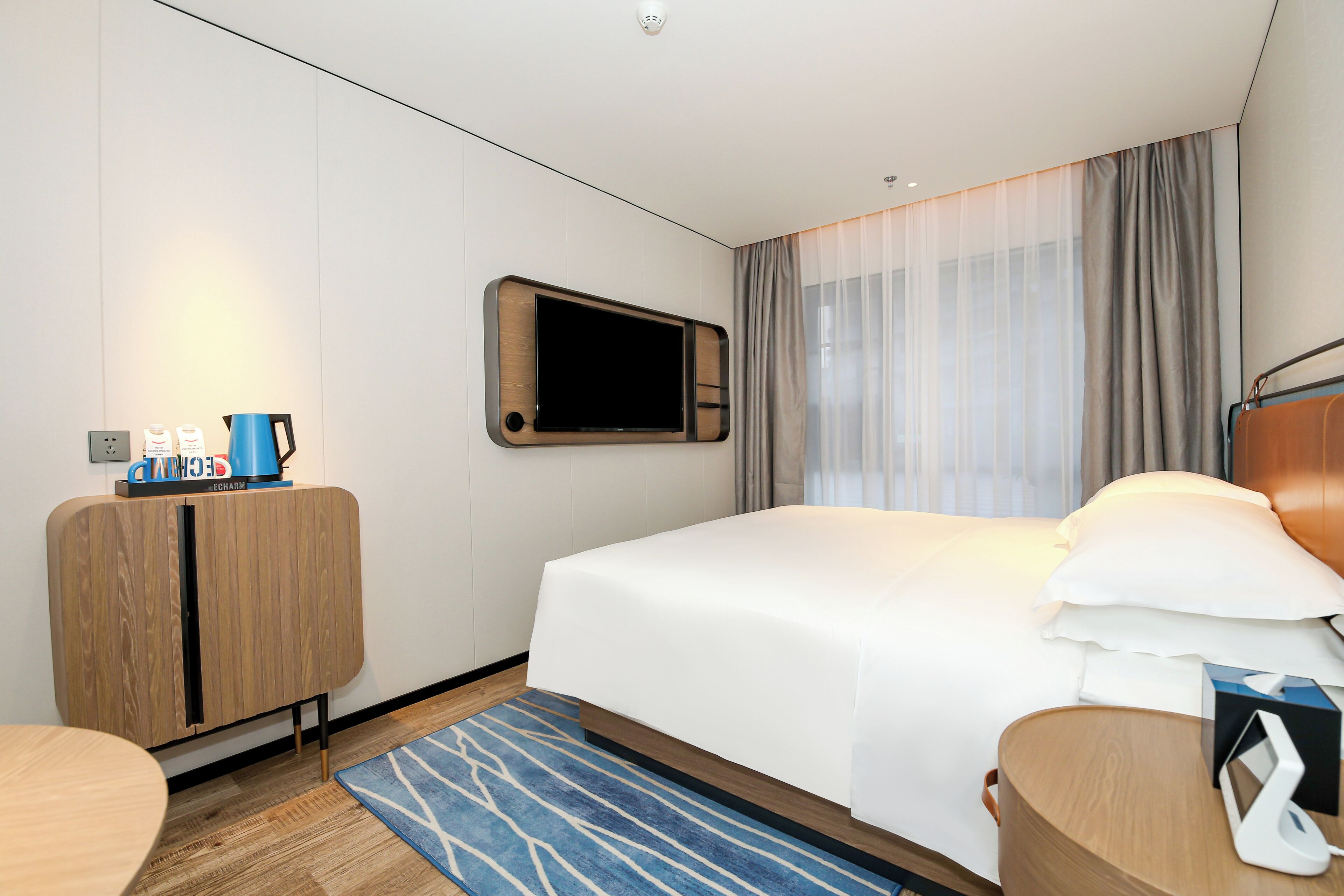 Quarto de hotel com cama, televisão fixada na parede, armário de madeira, escrivaninha e tapete azul estampado.
