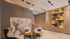 A modern lobby with a large abstract artwork, two white armchairs, a glass coffee table, and a wooden shelving unit with books and decorative items.