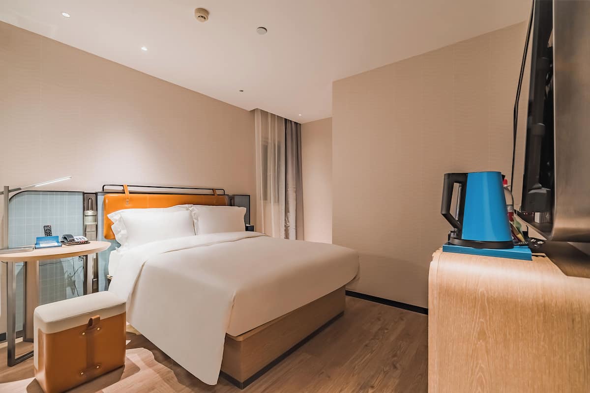 A hotel room with a bed, a desk with a lamp, a coffee machine, and a suitcase.