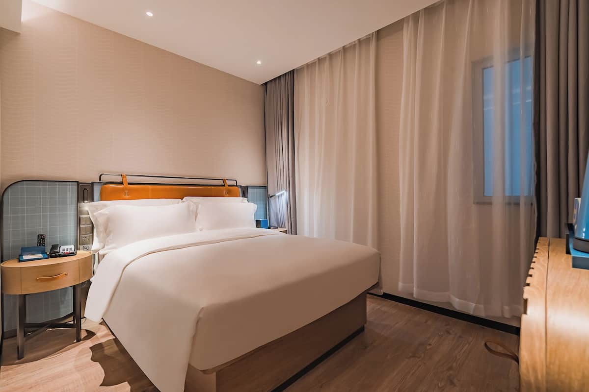 A hotel room with a large bed, a bedside table with a lamp, a wooden desk, and a window with sheer curtains.
