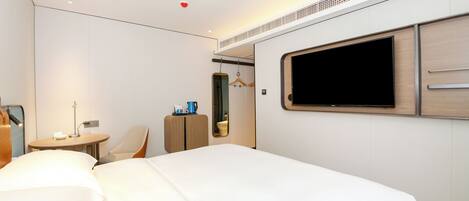 A hotel room with a large bed, a TV mounted on the wall, a small table with a lamp, and a wardrobe.
