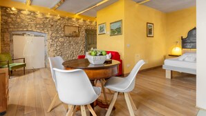 Uma sala de jantar com parede de pedra, mesa redonda de madeira e cadeiras brancas. Na sala adjacente, há uma cama com cabeceira.