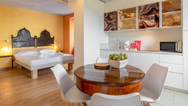 Quarto de hotel moderno com uma cama, uma mesa de madeira redonda, cadeiras brancas e uma área de cozinha com micro-ondas, além de arte decorativa nas paredes.