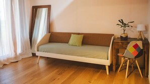 Una habitación acogedora con suelo de madera, un sofá con un cojín verde, una mesita de noche de madera con una lámpara, y una silla con respaldo colorido.