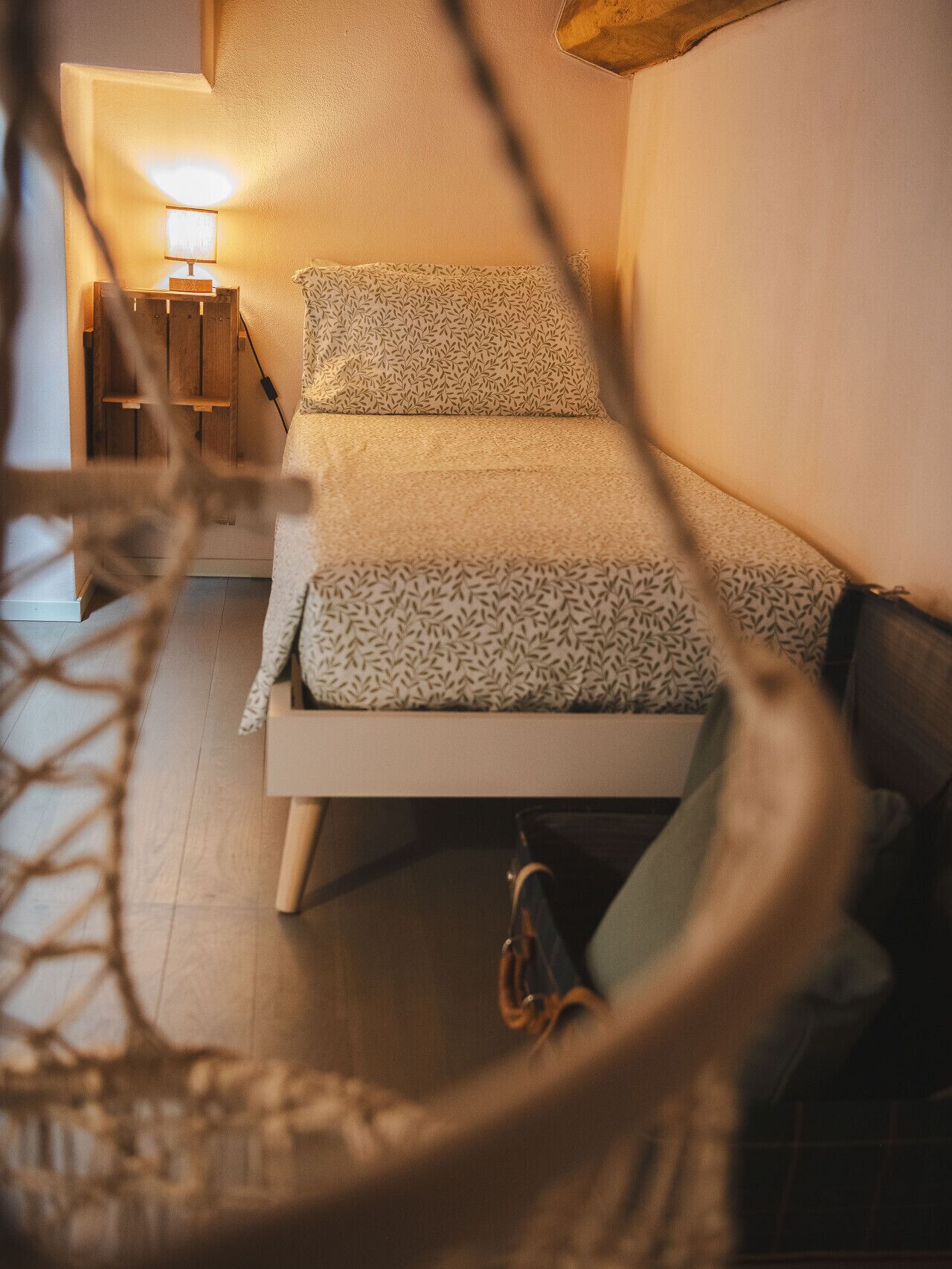 A bed with a patterned bedspread, a bedside table with a lamp, and a hanging chair in a room with wooden flooring.