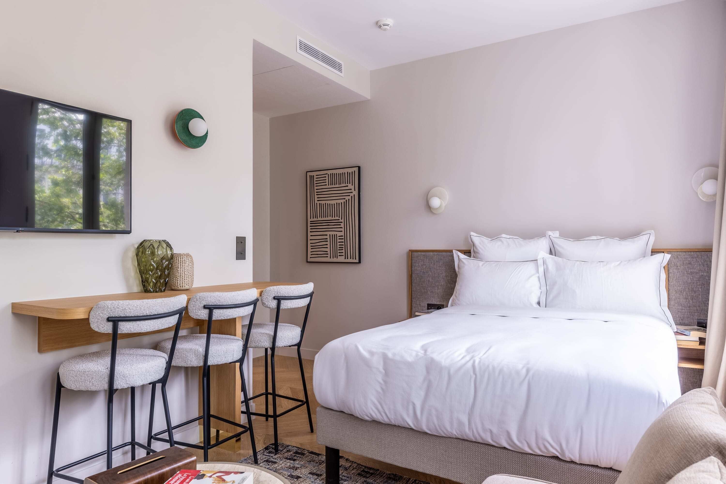 A modern hotel room with a large bed, a desk with bar stools, a flat-screen TV, and a decorative wall piece.