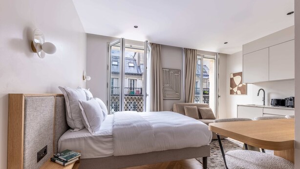 A modern hotel room with a bed, bedside table, books, a dining area, and a view of the city through large windows. - Marais - Studio in Paris (Paris)