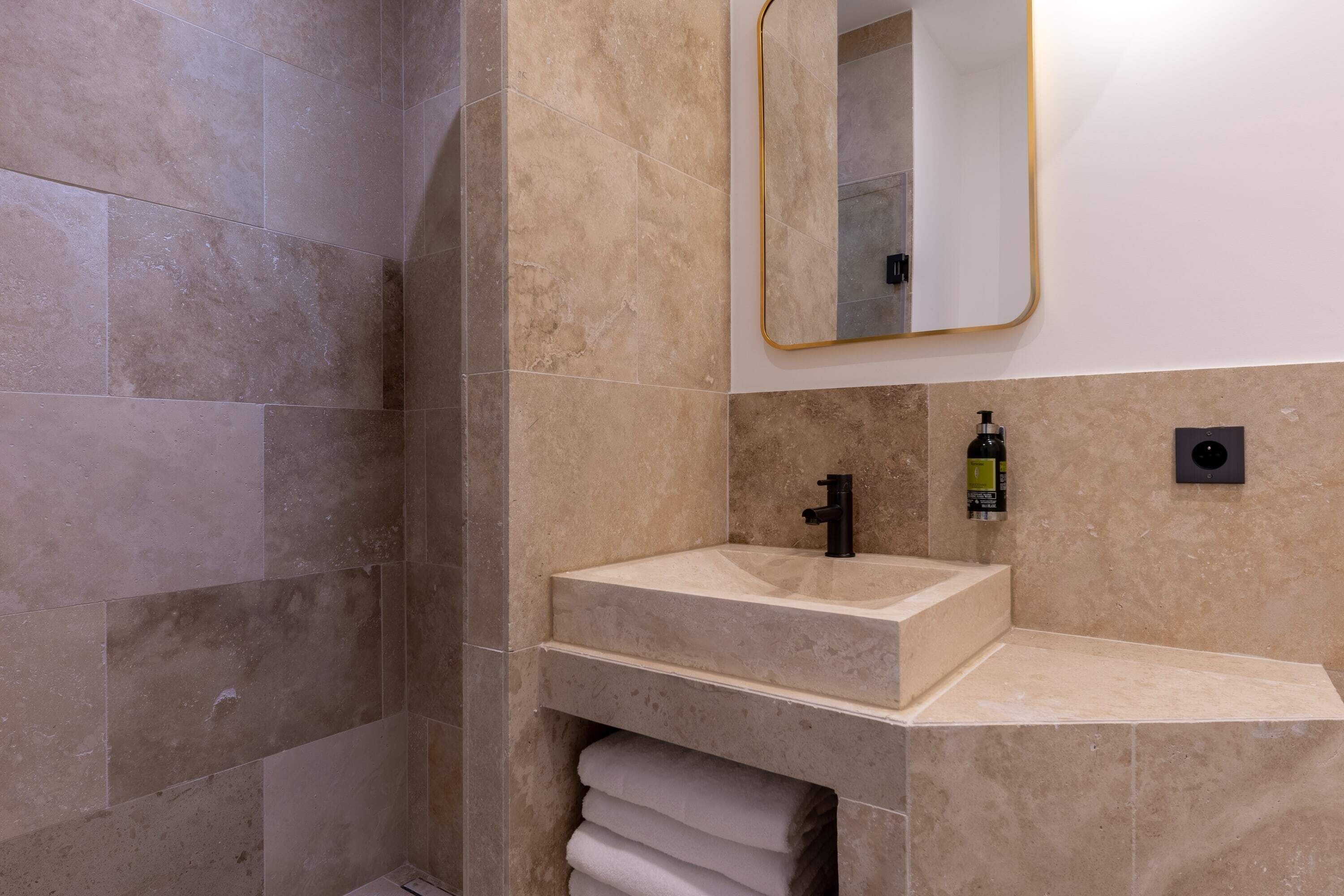 A bathroom with a beige tiled shower, a sink with a black faucet, and a mirror with a gold frame.