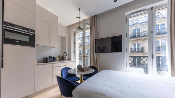 A modern hotel room with a bed, a dining table with chairs, a kitchen area, and a balcony with a view of the city.