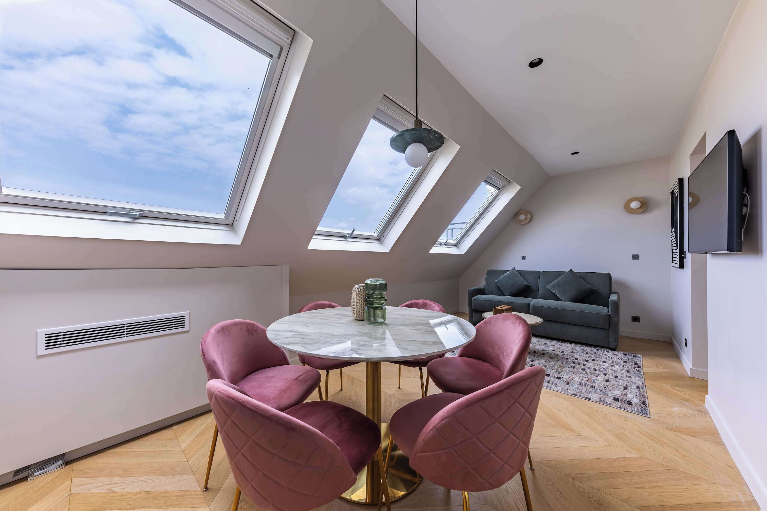 A modern living room with a round table, pink chairs, a grey sofa, and skylights.