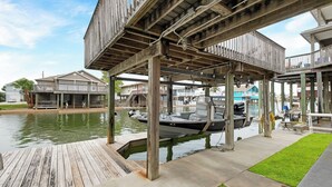 A wooden dock extending over a body of water with boats docked, a grassy area, and buildings in the background.