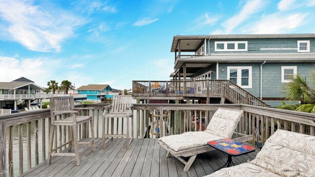 A wooden deck with lounge chairs and a small table, overlooking a body of water and a building with multiple levels.