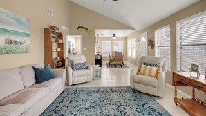 A spacious living room with a beige sofa, two armchairs, a coffee table, and a blue patterned rug. A ceiling fan and a wooden bookshelf are also visible.