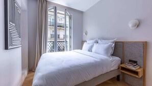A modern bedroom with a large bed, a nightstand, and a window with a view of buildings. - Marais - 2 Bedrooms in Paris (Paris)