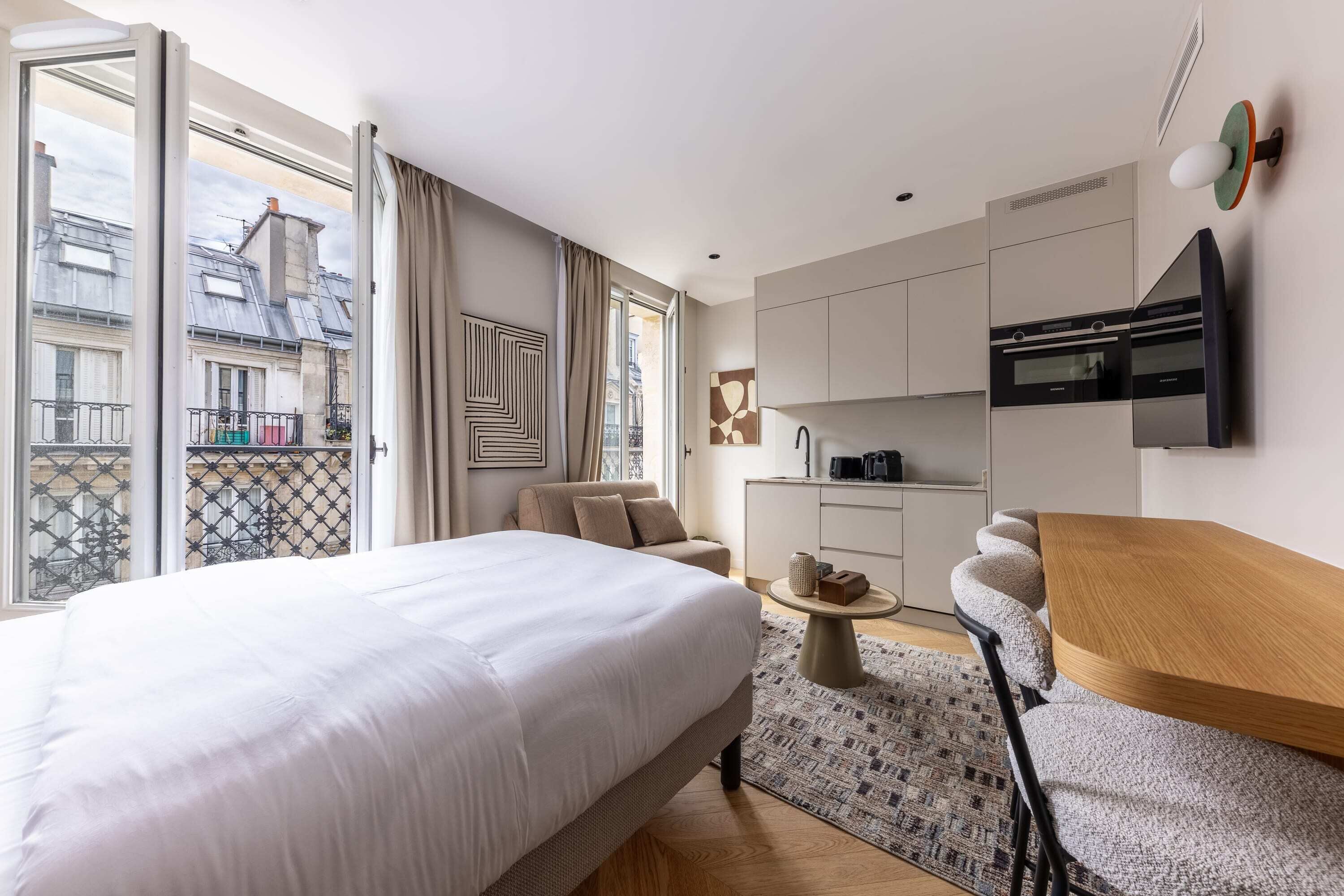 A modern hotel room with a large bed, a sofa, a dining table, and a kitchen area.